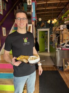 A male Moque-Tortue server holding a black plate with a flatbread and a bowl of broccoli soup at the foot of the bistro's colourful stairs in Shediac. He is wearing a black staff T-shirt with the turtle logo.