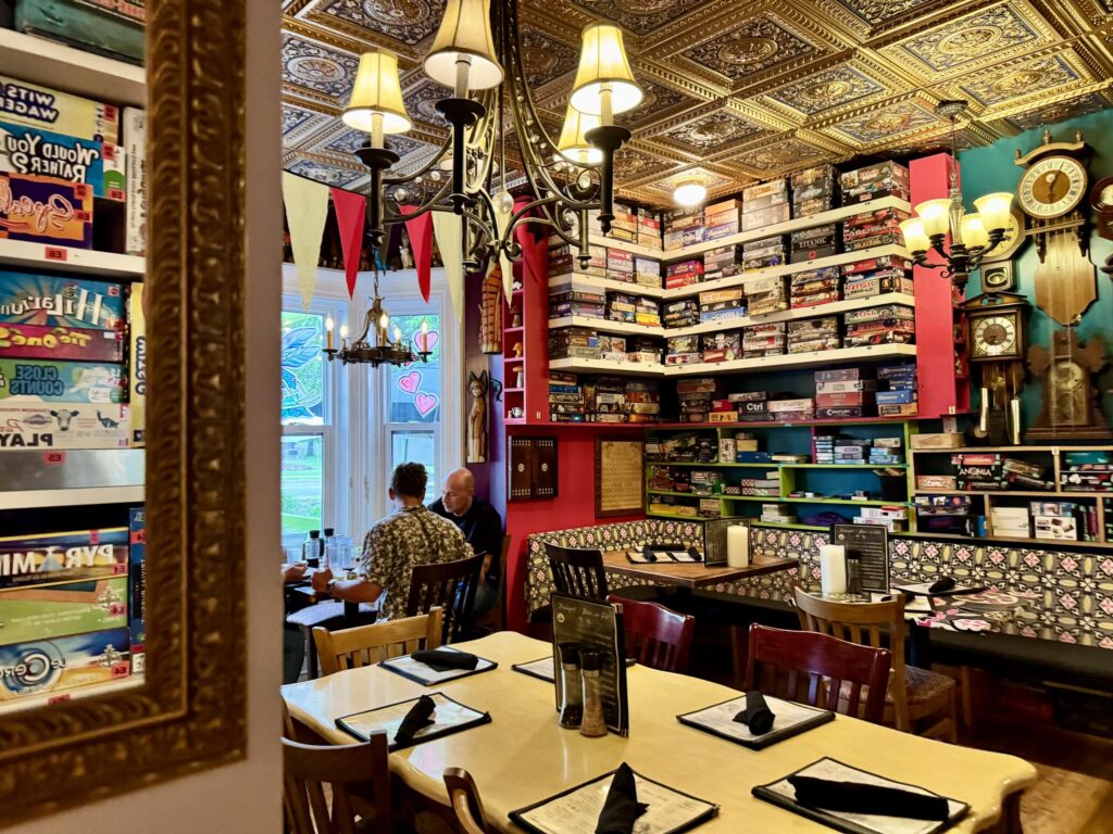 A group of three people sit at the window table in an otherwise empty dining room at Le Moque-Tortue. On the walls are clocks, mirrors, and hundreds of board games.