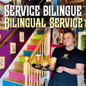 A smiling server at the bottom of Le Moque-Tortue's very colourful stairs carrying a tray of various beers and other drinks. She is wearing a black staff T-shirt